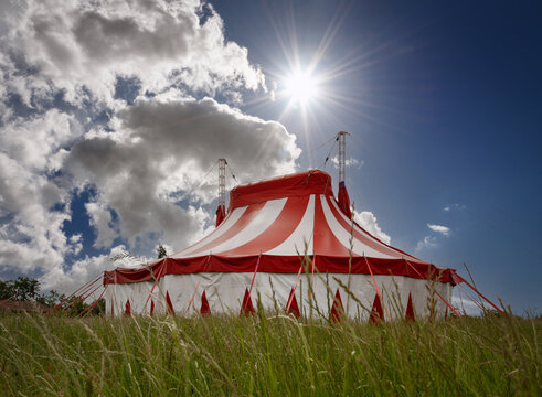Colourful circus tent on green meadow against a colorful sunset sky with sun rays and white clouds