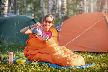 Woman in a sleeping bag drinks tea against the backdrop of many tents in a camping site at early...