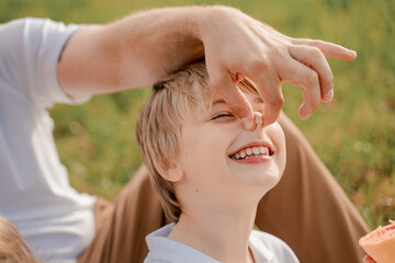Father plays with his son pinching his nose during family walk through meadow. International...