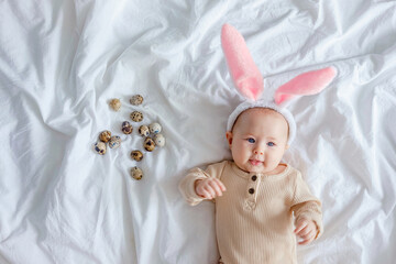 Fototapeta na wymiar An infant looks at the camera. Easter costume for children. A cute funny baby with bunny ears costume dressed up for Easter laying on white bed linen sheet with quail eggs.