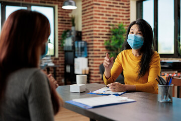 Asian entrepreneur with face mask against coronavirus discussing business partnership with freelancer woman working at management project in startup office. Executive managers collaborating together