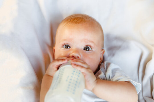 Cute little newborn girl drinking milk from bottle and looking at camera on white background. Infant baby sucking eating milk nutrition lying down on crib bed at home. Motherhood happy child concept
