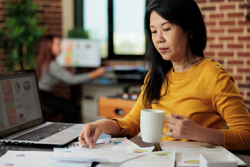 Asian businesswoman holding paper with management statistics working at business strategy in startup office. Executive manager analyzing graphs, brainstorming ideas for company increase