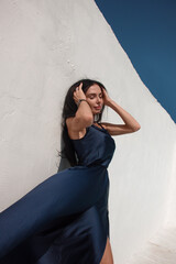 Fototapeta premium vacation mood. brunette woman in blue long silk dress is standing white wall background with sky at sunny day with hands near long hair and smiling. greek villa. travel concept, free space