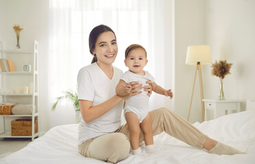 Portrait of a happy young mother together with her child at home. Beautiful mom sitting on bed and holding her cute baby wearing a diaper and comfy white cotton onesies. Family, care, comfort concept