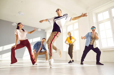 Expressive contemporary hip hop dance. Young male dancer performs hip hop movement on one leg during break dance lesson. Guy dances on surrounded by his dance friends in bright hall of dance studio.