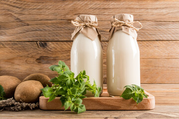 two glass bottles of vegetable potato milk on a wooden tray and a rustic table with tubers and...