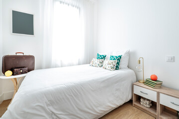 King size bed with white and green cushions, lamp without lampshade on bedside table and vintage brown travel suitcase