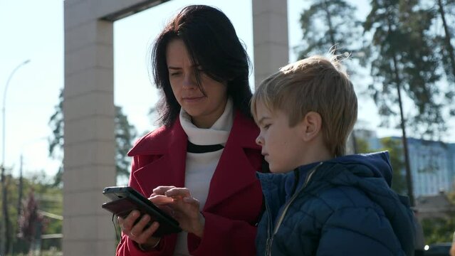 Mother and Son Watch Media on Phone. Woman Surfing Browsing Internet Using Smartphone. Mom with Kid rest in City Park. Autumn Day. 2x Slow motion 60fps 4K