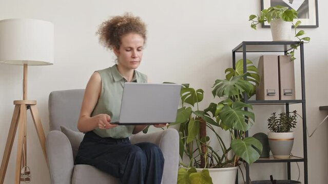 Medium slowmo of young Caucasian businesswoman sitting down in armchair with laptop on her laps while working from home