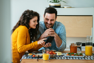 Happy Couple Using Mobile Phone Together While Eating Breakfast at Home