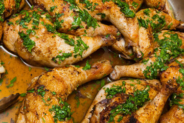 Baked chicken legs in own fat sprinkled with chopped parsley and garlic in a bowl with food tongs close-up.