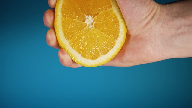 Close-up of a strong male hand squeezes juice from half of a juicy orange on a blue background