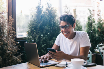 Cheerful male IT professional reading mobility notifications and smiling sitting at street coffee...
