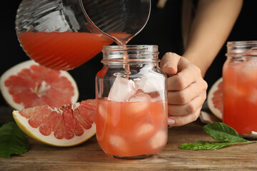 Woman pouring pink pomelo juice into glass jar with ice at wooden table, closeup