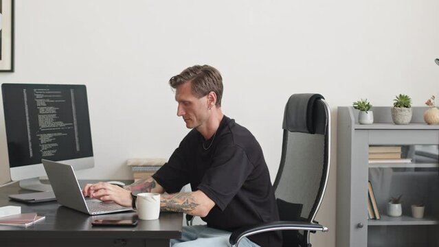PAN medium slowmo of young tattooed Caucasian software developer in casualwear sitting at desk in modern office typing program code on laptop connected to pc computer