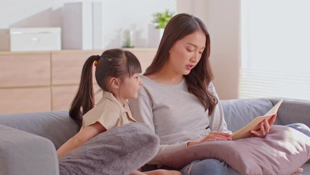 Mom and her daughter are sitting on the bland couch in the living room.Mother and kid reading together,the girl is really into her mom tale,she always smile while her mom has been reading.Mom and kid.