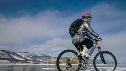 Woman is riding bicycle on the ice. Girl is dressed in a silvery down jacket, cycling backpack and helmet. Ice of the frozen Lake Baikal. Tires on bike are covered with spikes. Traveler is ride cycle.