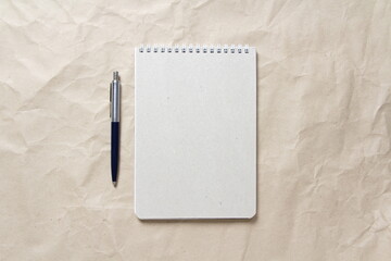 Gray notepad with white coiled spring and pen on a background of beige crumpled craft paper. With empty space for text and design