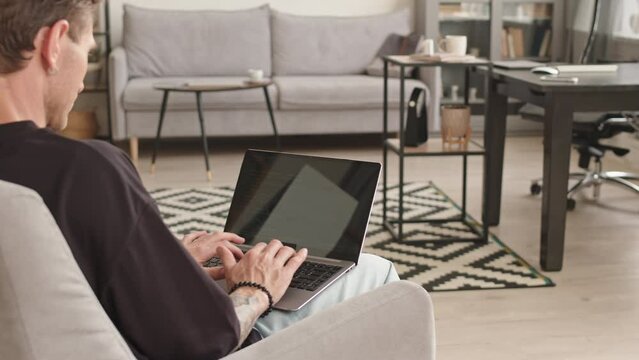 PAN rear view of Caucasian male programmer with tattooed arms typing program code on laptop sitting on couch at home office