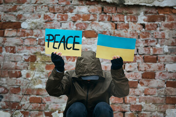 Unrecognizable person holds Ukrainian flags with peace message against war damaged wall.