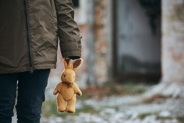 Unrecognizable refugee with plush bunny on war damaged city street.