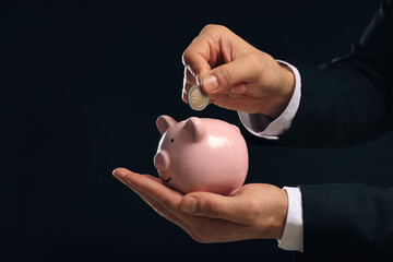 Businessman putting coin in piggy bank on black background, closeup