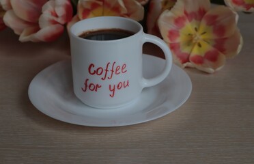 A white mug with black coffee stands against a background of tulips on a light surface.