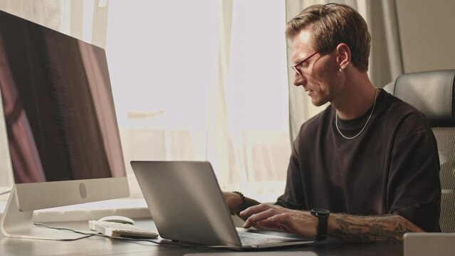Medium shot of young Caucasian male programmer typing on laptop sitting at desk by window on sunny day