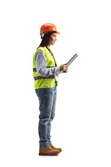 Full length profile shot of a young female engineer with a safety vest and hardhat holding a clipboard