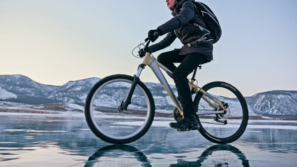 Fototapeta na wymiar Man is riding bicycle on the ice. Ice of the frozen Lake Baikal. Teenage is dressed in black down jacket, cycling backpack, helmet. Tires on covered with special spikes. Traveler boy is ride cycle.