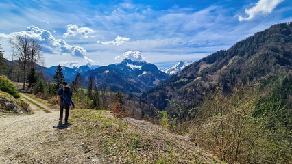 Fototapeta na wymiar Man with backpack and scenic view of snow capped mountain peaks of Karawanks near Sinacher Gupf in Carinthia, Austria. Mount Hochstuhl (Stol) visible through forest in spring. Rosental sunny day. Awe