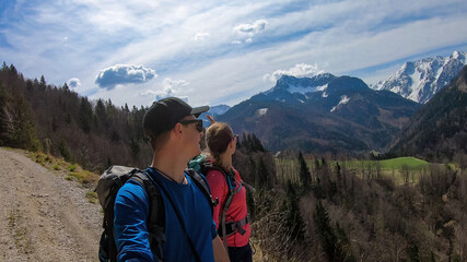 Obraz premium Couple with backpack and scenic view of snow capped mountain peaks of Karawanks near Sinacher Gupf in Carinthia, Austria. Mount Hochstuhl (Stol) visible through forest in spring. Rosental sunny day