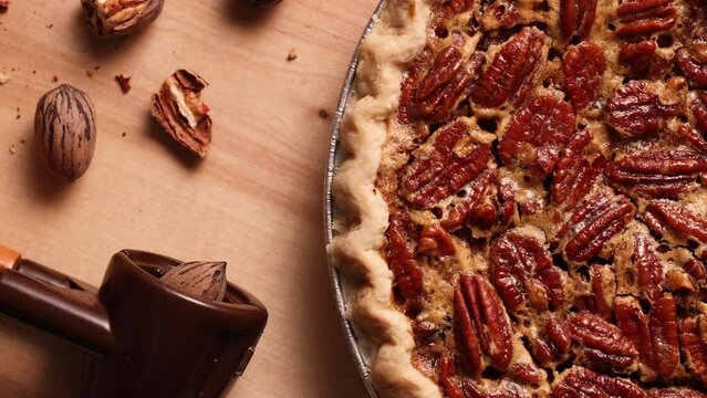 Revealing shot of pecan pie appearing in frame. Pecan pie being placed on wooden table with raw nuts and nutcracker, top view.