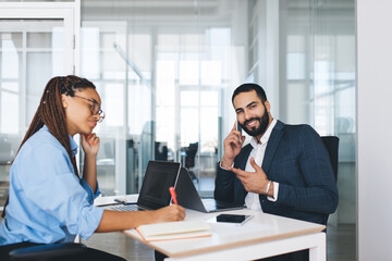 Blurred female employee writing text idea for business startup project while cheerful male boss using laptop smartphone technology for calling, happy corporate director smiling at camera while phoning