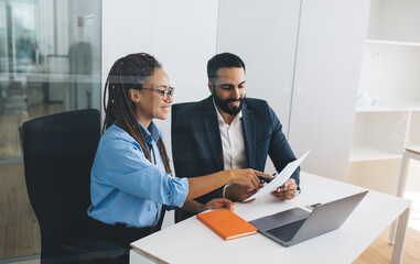 Multiracial male and female professional economists analyzing report during work time in office interior, happy colleagues discussing ideas for business and startup project using paperwork and netbook