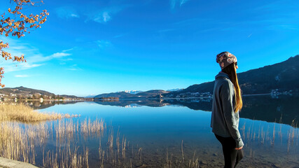 Fototapeta premium Happy woman walking along the shore of beautiful Woerthersee in Poertschach, Carinthia, Austria. Scenic lake landscape surrounded by Alps. Serenity. Fresh and clean air. Reflection in Lake Woerth