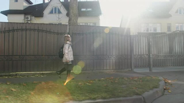 Blonde-haired school-age girl in a pink jacket with a backpack, walking home on a sunny fall or spring day. Steadicam shot, side view