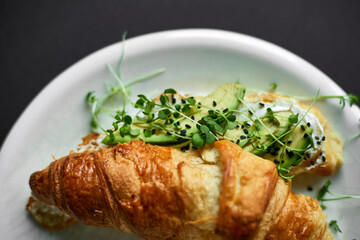 Croissant sandwich with cream cheese avocado served with micro green on plate black background.