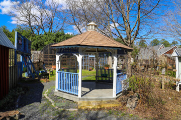 a white, blue and brown gazebo in the garden surrounded by bare winter trees and lush green plants...