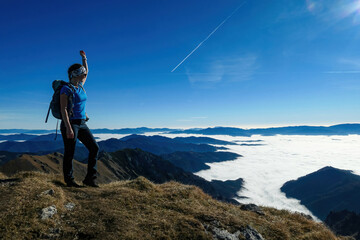 Obraz premium Woman with backpack hiking and admiring a scenic view from mount Eisenerzer Reichenstein in Styria, Austria, Europe. Ennstal valley is covered in clouds. Hiking trail, Wanderlust. Freedom concept