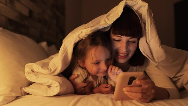 Pretty mom relaxing with little girl child using cellphone together, while lying on bed under the blanket. Happy young mother resting with small daughter watching learning video on smartphone