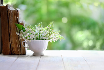 Lily of the valley in cup and books on white table, green natural background. symbol of spring...