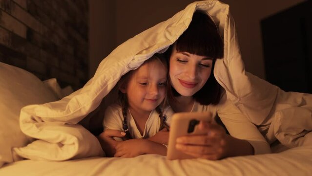 Pretty mom relaxing with little girl child using cellphone together, while lying on bed under the blanket. Happy young mother resting with small daughter watching learning video on smartphone
