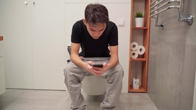 A young man sits on the toilet with a smartphone in his hands, writes a message.