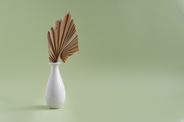 Ceramic white vase with a palm leaf on a dull green background. Minimalistic composition of a vase on a colored background with a place for an inscription.