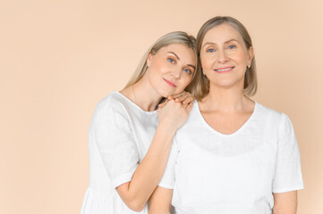 Mom and daughter on a beige background. Portrait of a young woman and her old charming mother. White clothes.