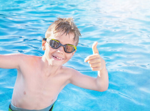 Healthy lifestyle. Active, happy nine years old child (boy) in sport goggles showing thumbs up on the swimming pool background. Caucasian kid learning to swim and enjoying water. Swimming activities.