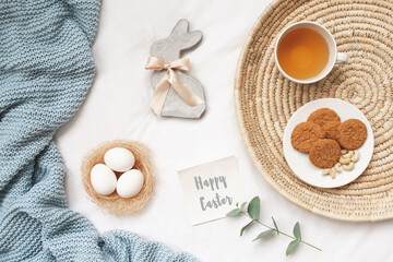 Fototapeta na wymiar Wicker tray, nest with white eggs, rabbit made of concrete, cup of tea, cookies, blue knitted plaid or blanket and card with greeting Happy Easter. Easter decor. Flat lay, top view.