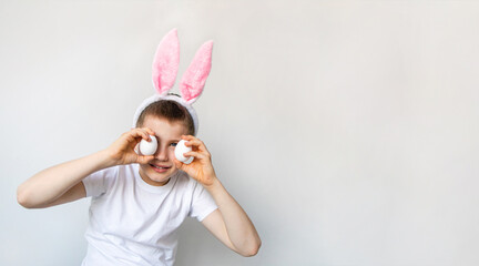 Banner of playful boy with bunny ears keeping white eggs near eyes on white background. Happy child boy is wearing bunny ears and white t shirt.Preparing for the holiday is a joy for kids. 
